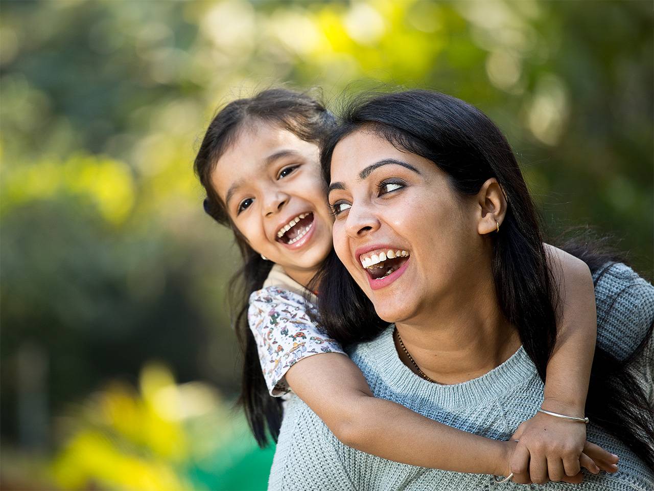 Woman with long black hair laughs while carrying her smiling daughter on her back outdoors