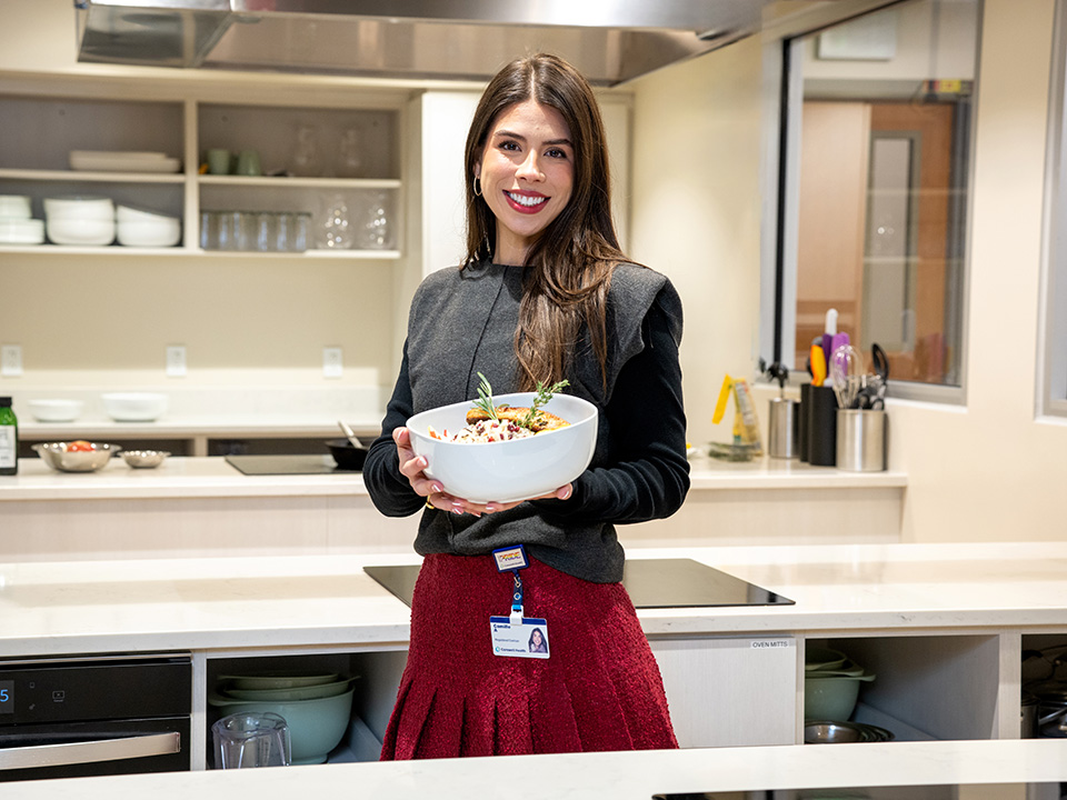 A woman in a kitchen holds a bowl of food, showcasing her culinary preparation with a cheerful demeanor.