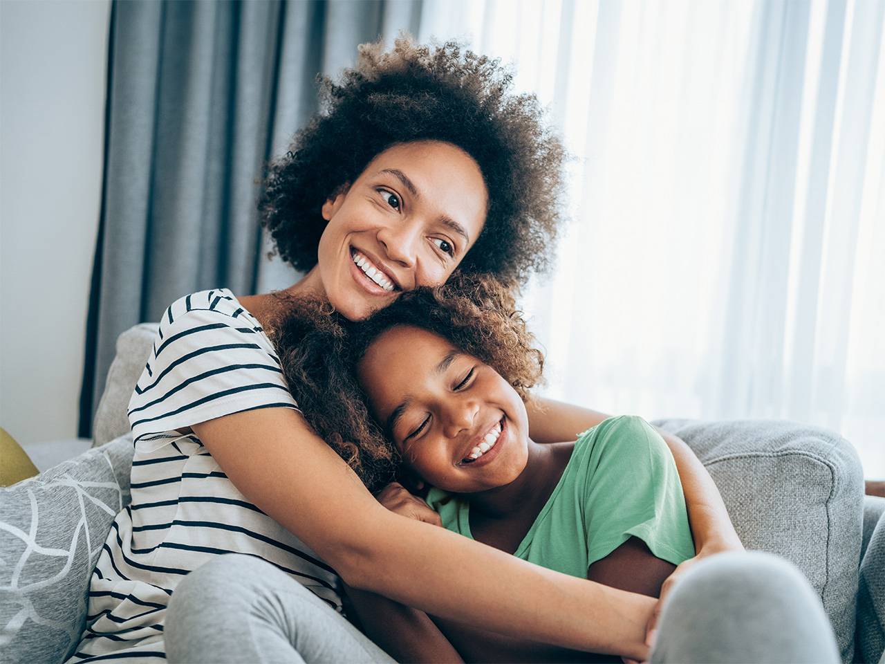 Smiling Black woman wearing a white shirt with navy blue stripes sits on a couch and hugs her smiling daughter