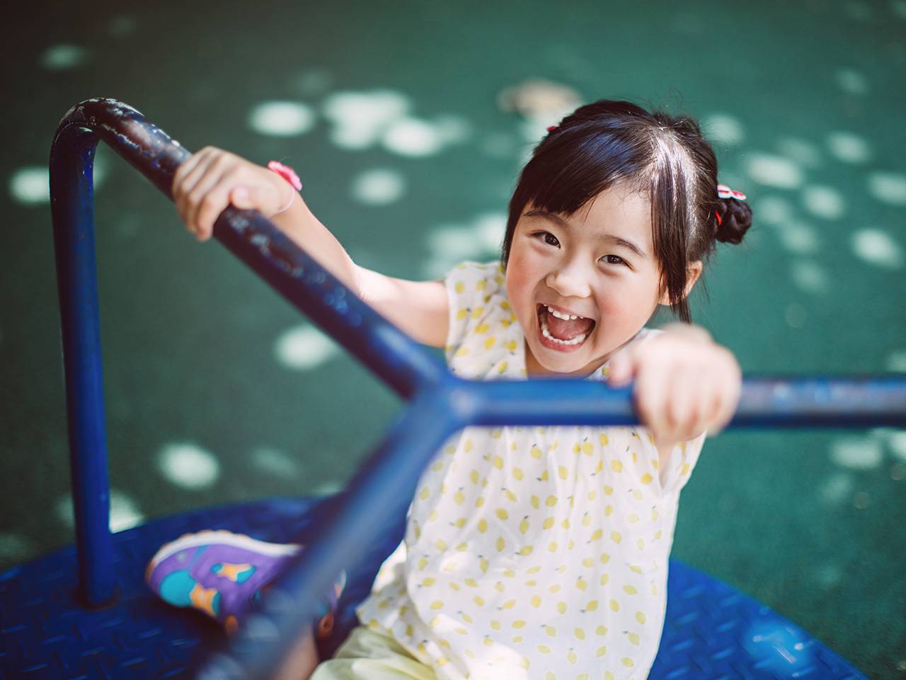 Girl with black hair and wearing a white shirt with yellow polkadots smiles and laughs while riding on a merry go round