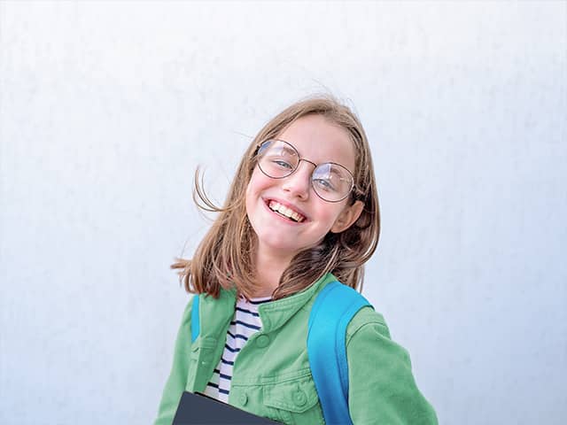 Preteen girl wearing a green shirt and glasses stands in front of a white wall outdoors