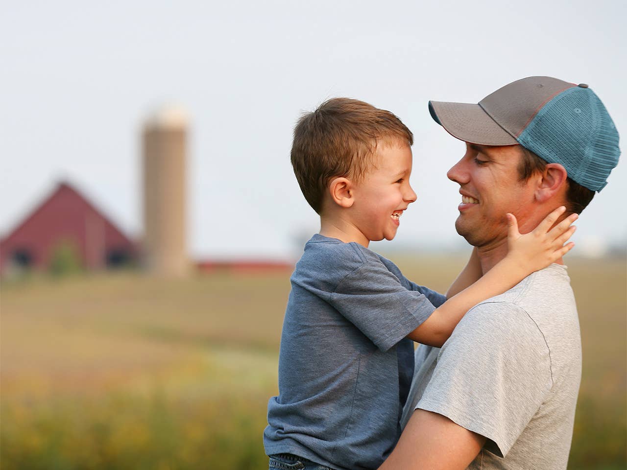 Smiling young white man lifts up his young son while they stand in a field with a barn and silo in the background