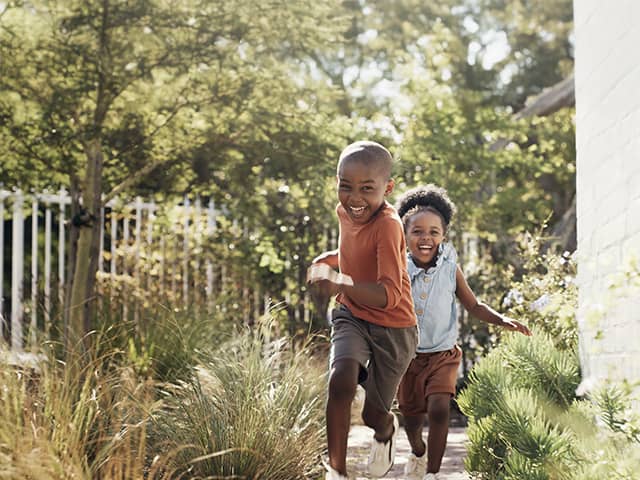 Smiling young Black boy and his smiling sister run together through tall wild grass in the sunshine outdoors