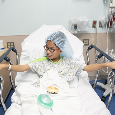 Smiling young boy with glasses in surgical gown sitting in hospital bed