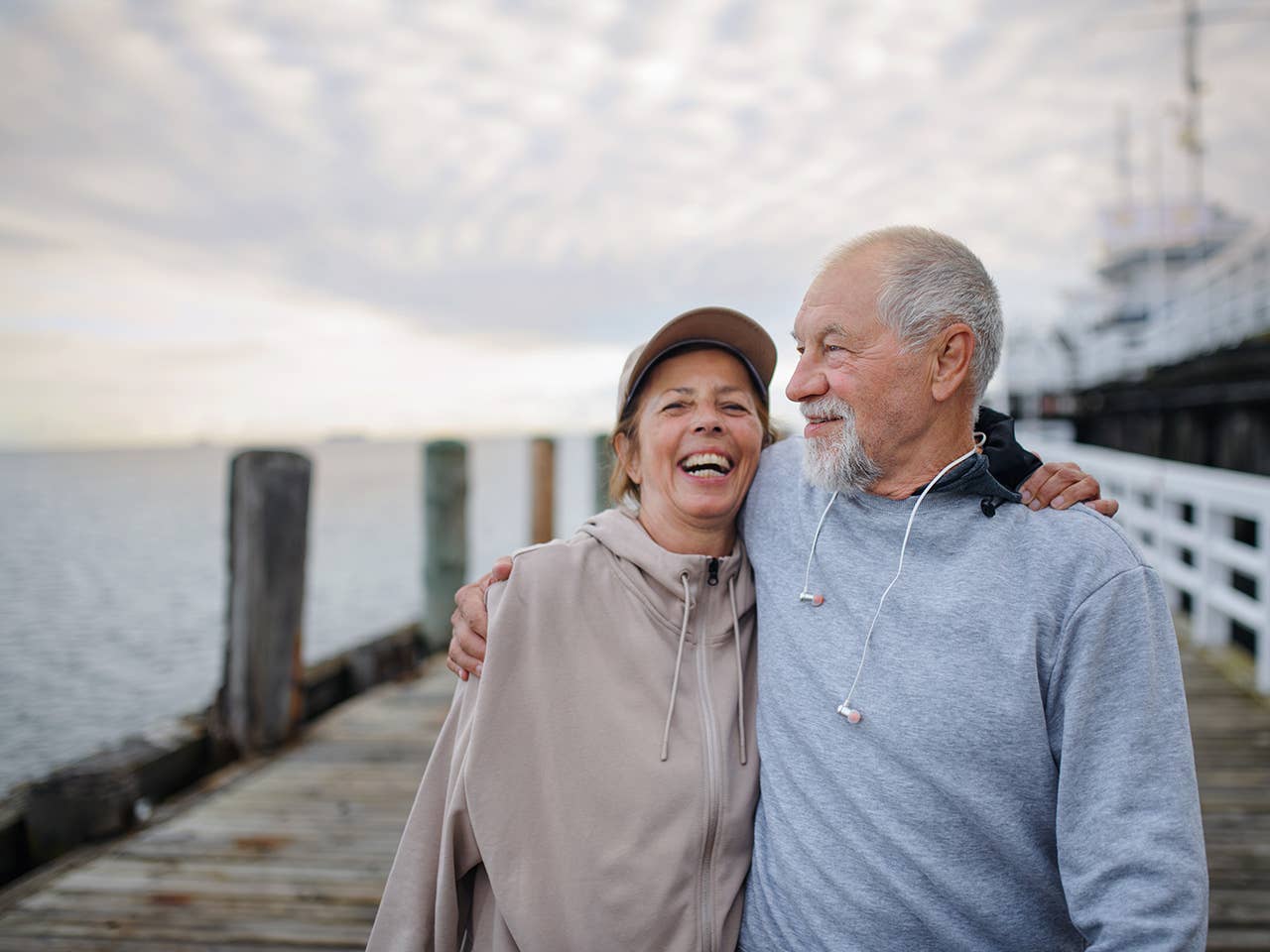 Laughing woman in a tan zip-up sweatshirt hugs a man while standing outdoors on a dock