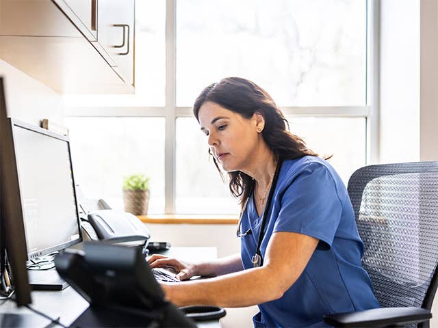 A woman in scrubs seated at a desk, focused on her work while using a computer.