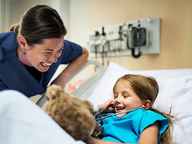 Female medical professional smiling and watching a young female patient using a stethoscope on her toy bear while lying in a hospital bed