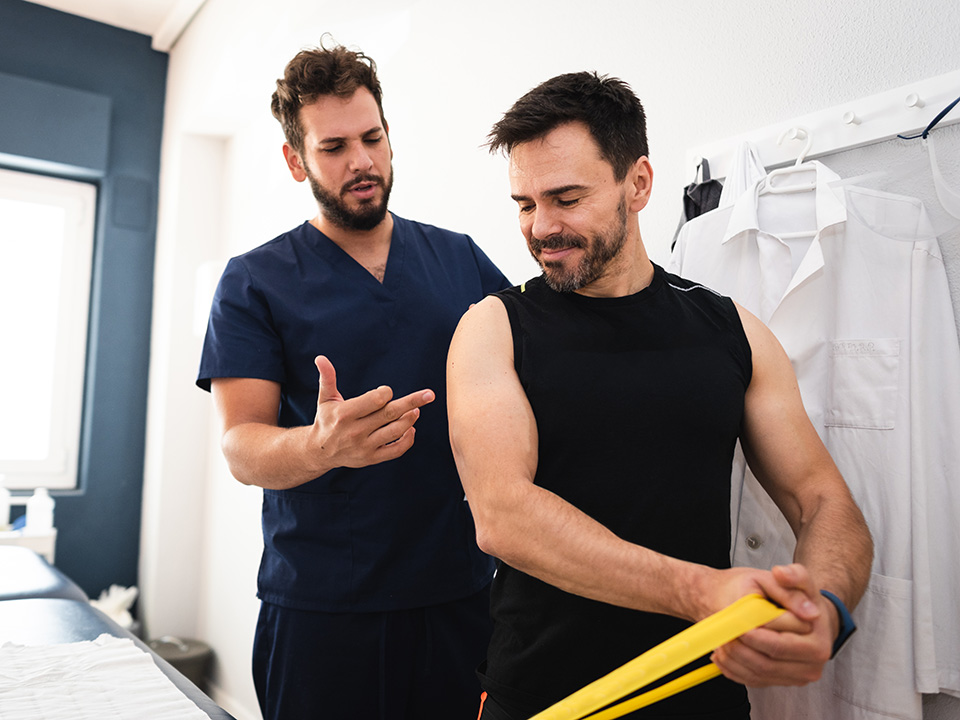 Middle aged white man stretches using an exercise band while he is assisted by a Corewell Health Orthopedics@Work specialist