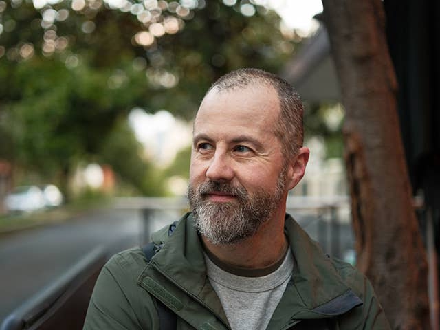 Older white male with balding grey hair and a beard stands outdoors as the evening sun drops below nearby trees