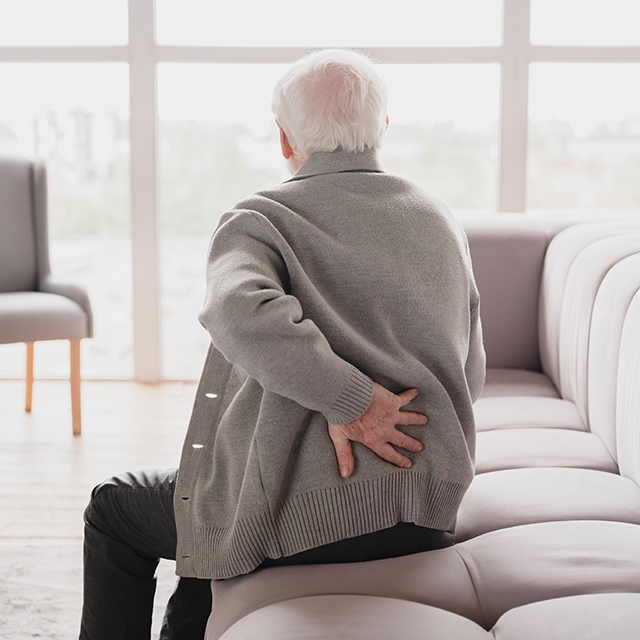 An older man sitting on a couch, visibly uncomfortable, holding his lower back due to pain.