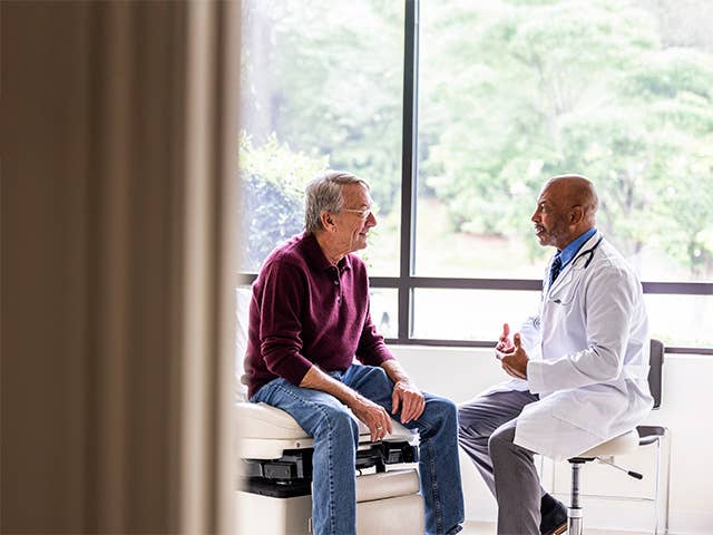 Elderly man wearing glasses and a maroon sweater sits on exam table speaking with male doctor