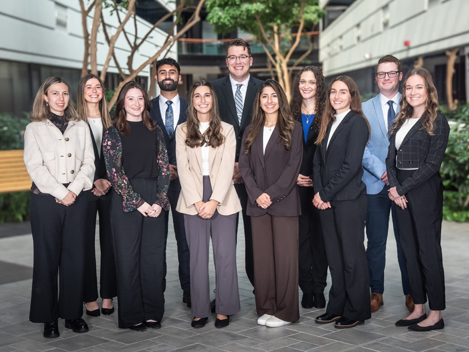 Group of 10 healthcare residency participants in professional attire standing in a bright indoor atrium.