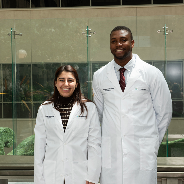 Two pediatric chief residents in white lab coats standing in a hospital atrium with glass walls and indoor landscaping.