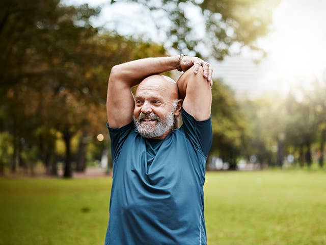 White man with a bald head and grey beard stretches his arms over his head while standing outdoors