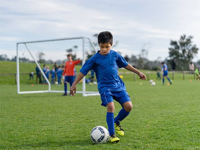 Boy with black hair and wearing a blue short sleeve shirt and blue shorts dribbles a soccer ball on a grassy soccer field outdoors
