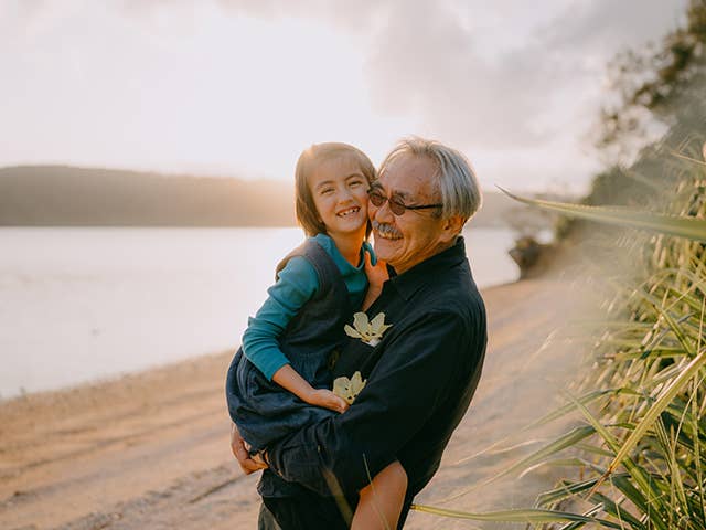 Elderly Asian man lifts up his granddaughter as they stand on a beach
