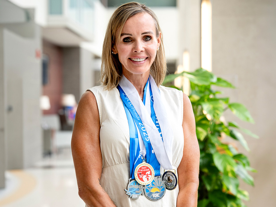 Christine Ankenbauer, a white woman with long blonde hair, wearing a white shirt and five running medals around her neck, standing in front of a hospital building