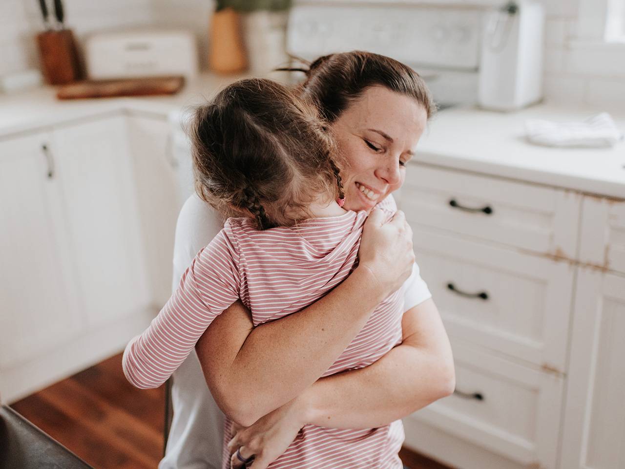 Smiling white woman kneels down and hugs her daughter in the kitchen