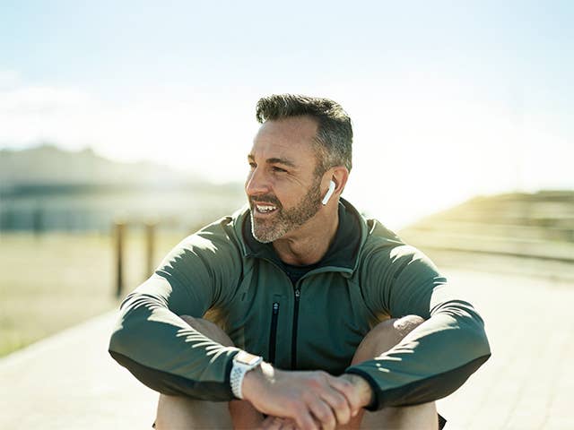 Man wearing a dark green jacket sits in the sunshine wearing earbuds