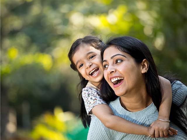 Woman with long black hair laughs while carrying her smiling daughter on her back outdoors