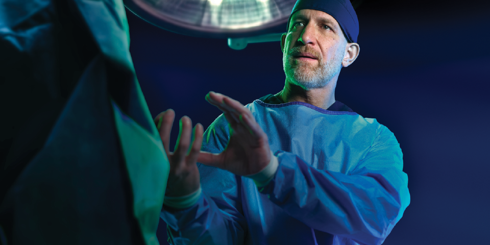 Healthcare professional in surgical attire gesturing with hands beneath an operating room light.