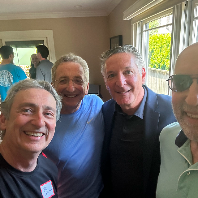 Four attendees pose together indoors at a social gathering, with additional guests visible in the background.