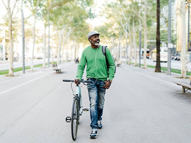 Middle aged Black man wearing a green sweater walks his bike through a park