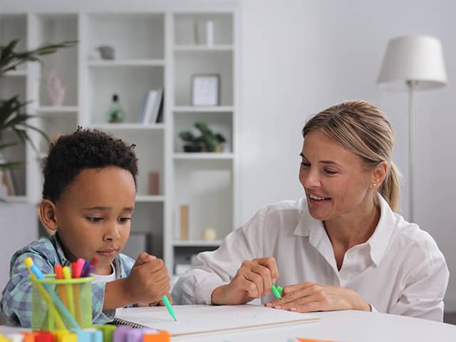Smiling woman wearing a white blouse sits next to a young Black boy at a table as they color with markers