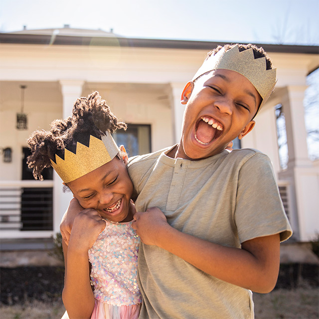 Smiling Black boy wearing a toy crown wraps his arm around his smiling sister as they play outdoors