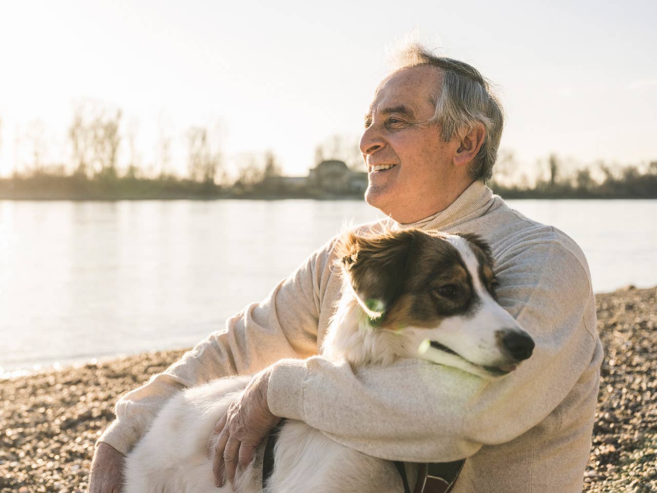 Older white man wearing a white sweater sits with a dog next to a small lake