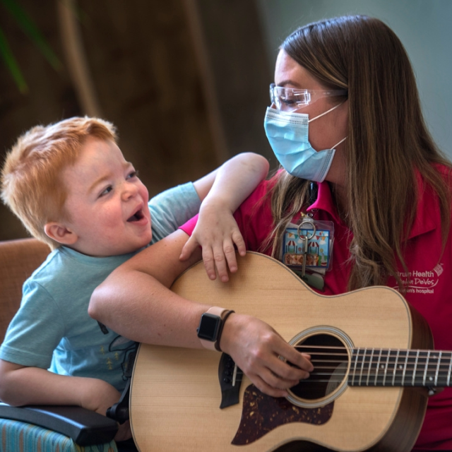 Woman in a burgundy shirt wearing a mask plays an accoustic guitar for a young boy