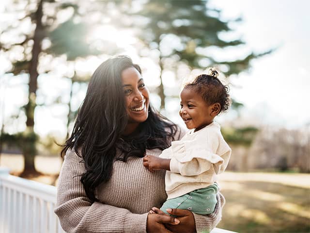 Smiling Black woman wearing a beige sweater holds her daughter as they stand outdoors in the sunshine