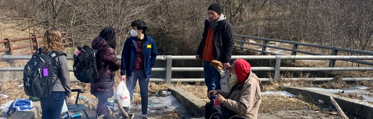 A diverse group of individuals stands on a bridge, each carrying bags, symbolizing community support and outreach efforts.
