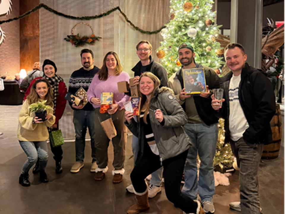 A group of people gathered indoors for a winter or holiday event, standing in front of a decorated Christmas tree and holding various gifts.