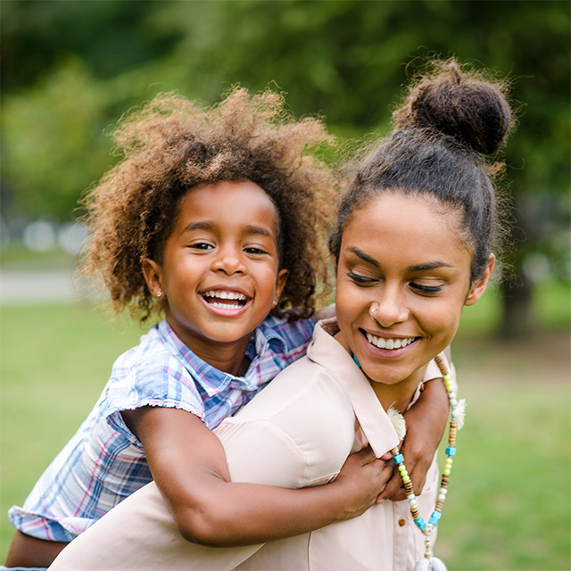 Smiling Black woman looks back at her smiling daughter while carrying her on her back outdoors