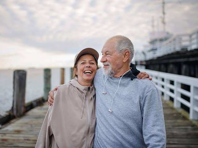 Laughing woman in a tan zip-up sweatshirt hugs a man while standing outdoors on a dock