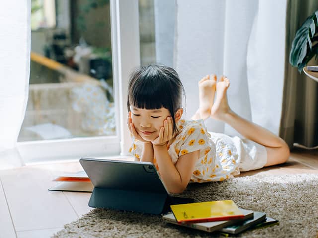 An Asian girl wearing a white shirt with yellow polka dots lies on the floor in a sunlit room looking at a tablet device