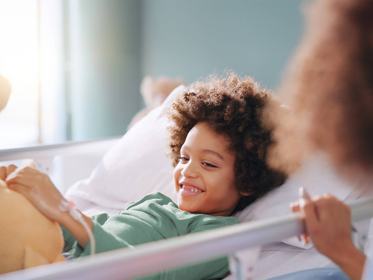 A young Black boy with curly brown hair lies in a bed inside a sunlit room