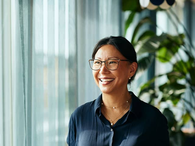A smiling woman wearing glasses stands in front of a window, with natural light illuminating her face.