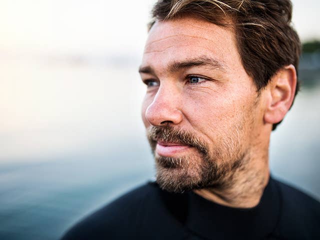 Man with brown hair and a goatee stands next to a lake, looking into the distance