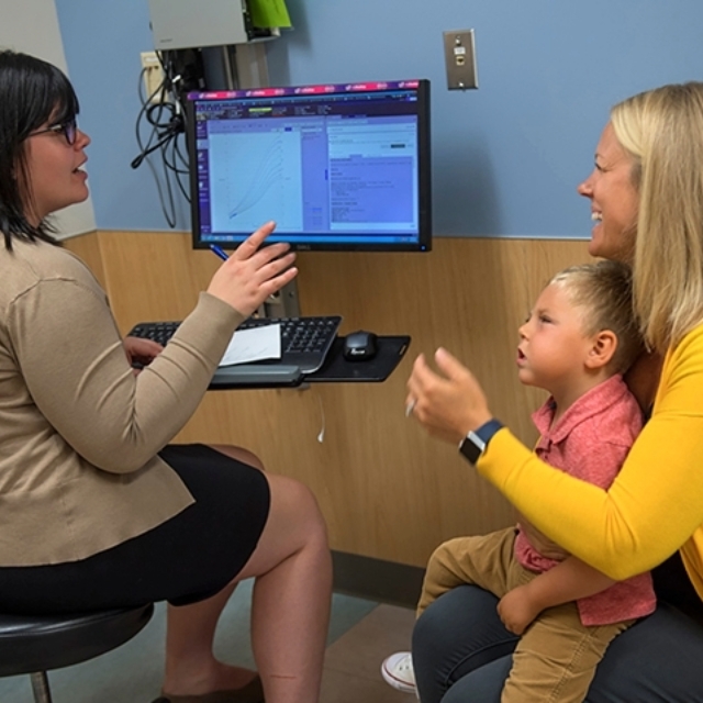 Smiling mother sits with young son in her lap, speaking to doctor in epilepsy clinic