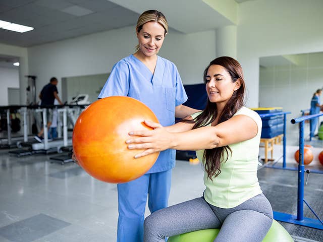 Therapist in light blue scrubs assists a woman holding an inflatable exercise ball in her outstretched arms