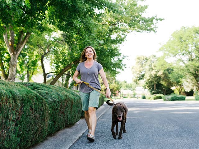 Smiling middle aged white woman walks a black dog in the shade of trees on a sunny day