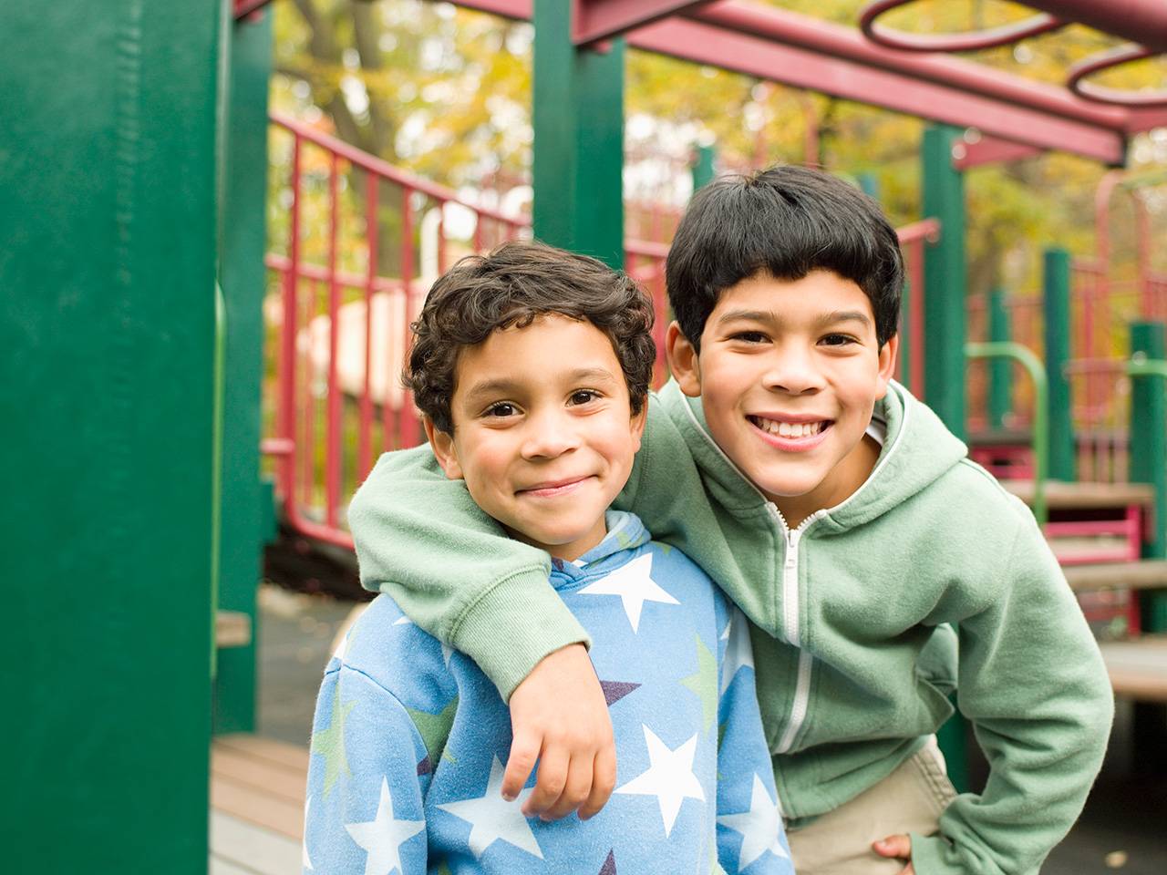 Smiling preteen boy with black hair stands with his arm around his younger brother's shoulder on a playground