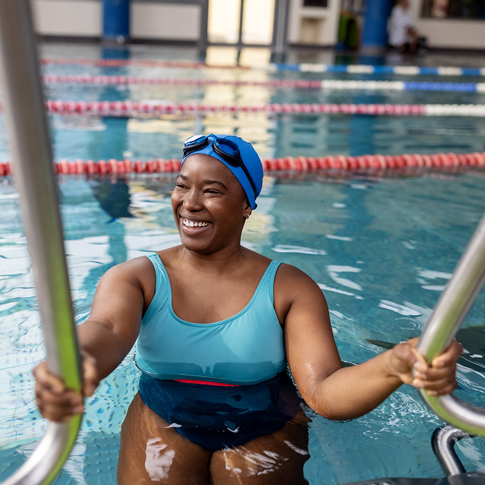 Smiling Black woman in a blue swimsuit stands on ladder as she climbs into swimming pool