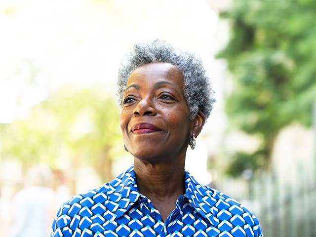 Elderly Black woman wearing a blue shirt with white polka dots stands outside in the sunshine