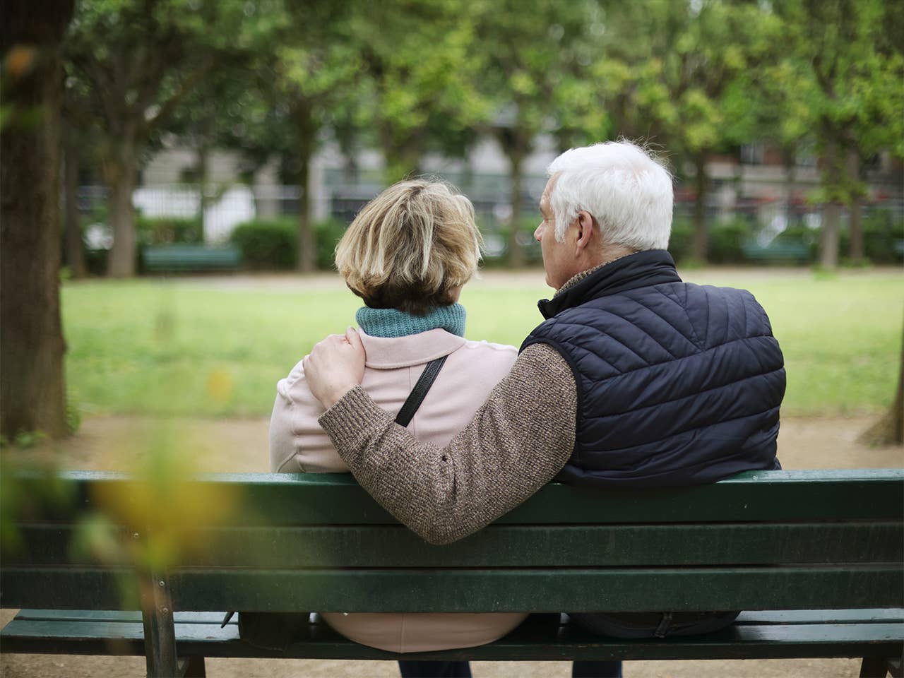 Elderly man sits next to his wife on a park bench, with a hand on her shoulder