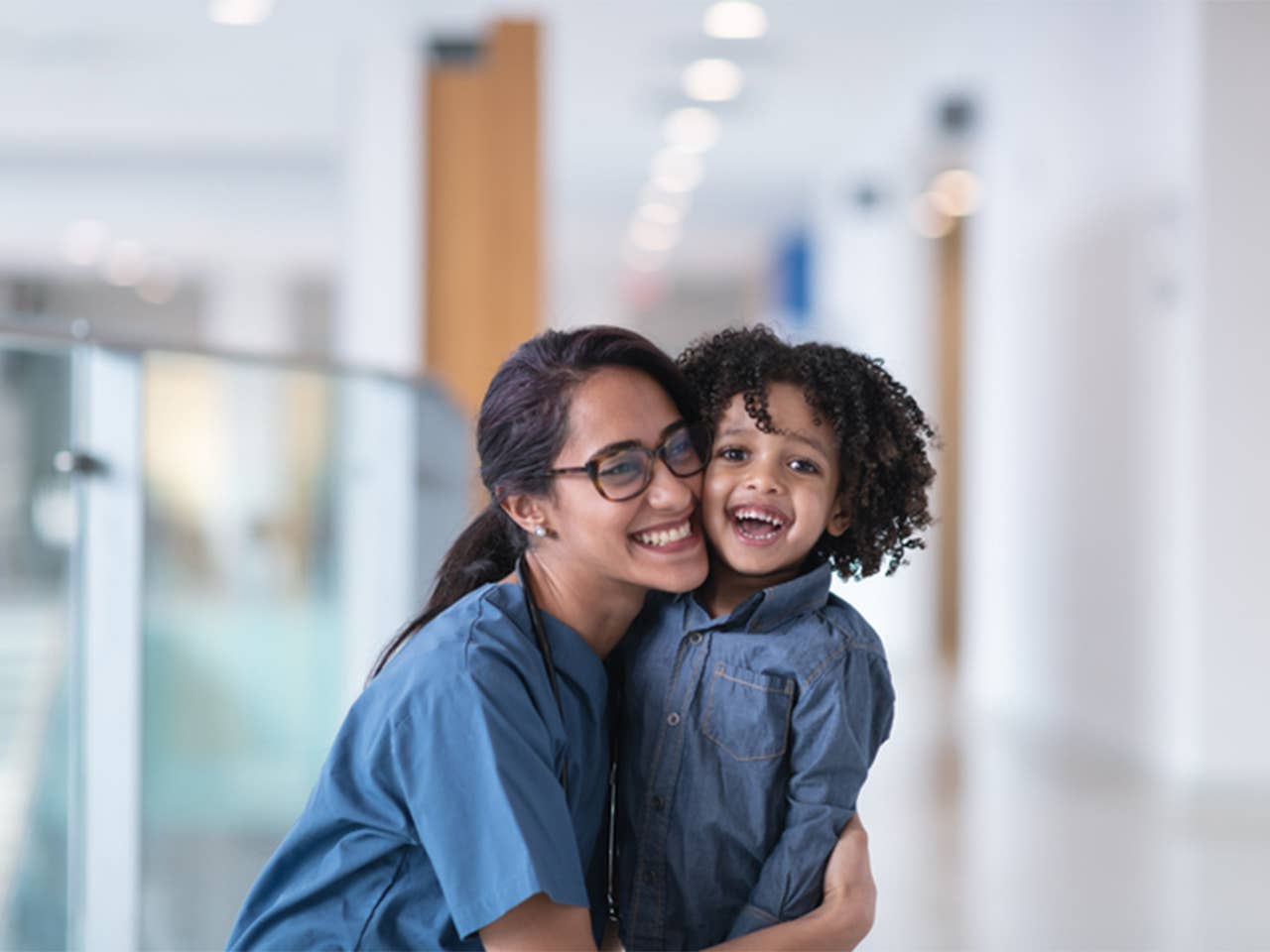 Woman in blue scrubs hugging a child in a hospital hallway