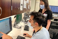 Two individuals in face masks engaged in computer work, representing a collaborative effort in Resident Teaching.