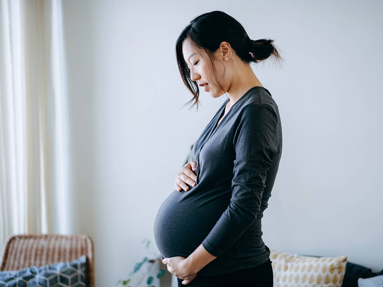 Pregnant Asian woman stands in a sunlit room with her hands on her belly